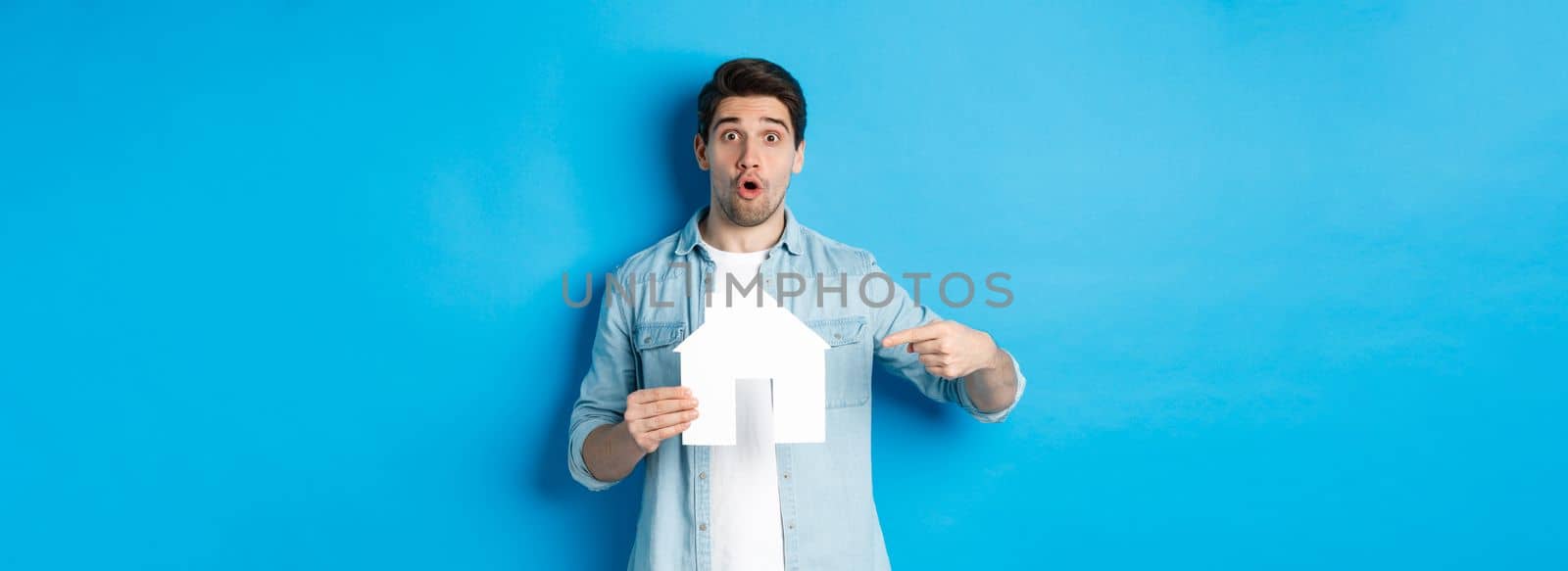 Insurance, mortgage and real estate concept. Surprised young man pointing at house card model and looking at camera, standing against blue background.