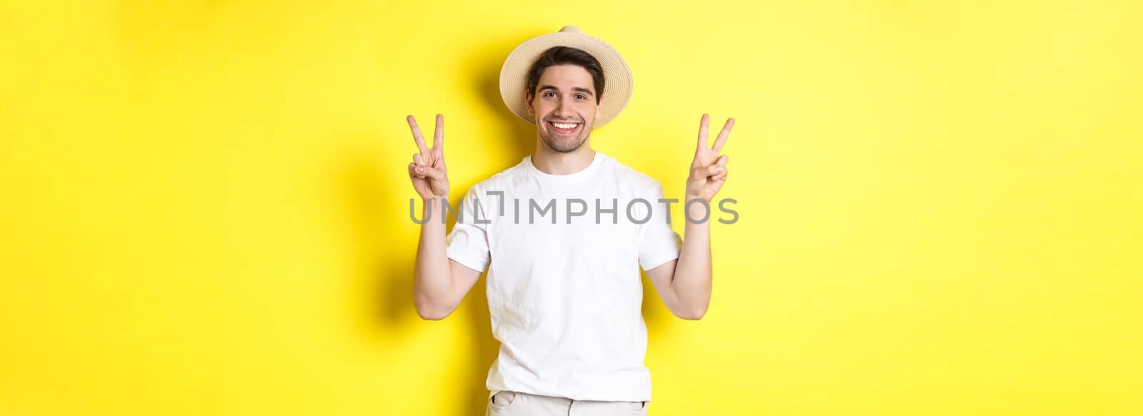 Concept of tourism and vacation. Happy male tourist posing for photo with peace signs, smiling excited, standing against yellow background.