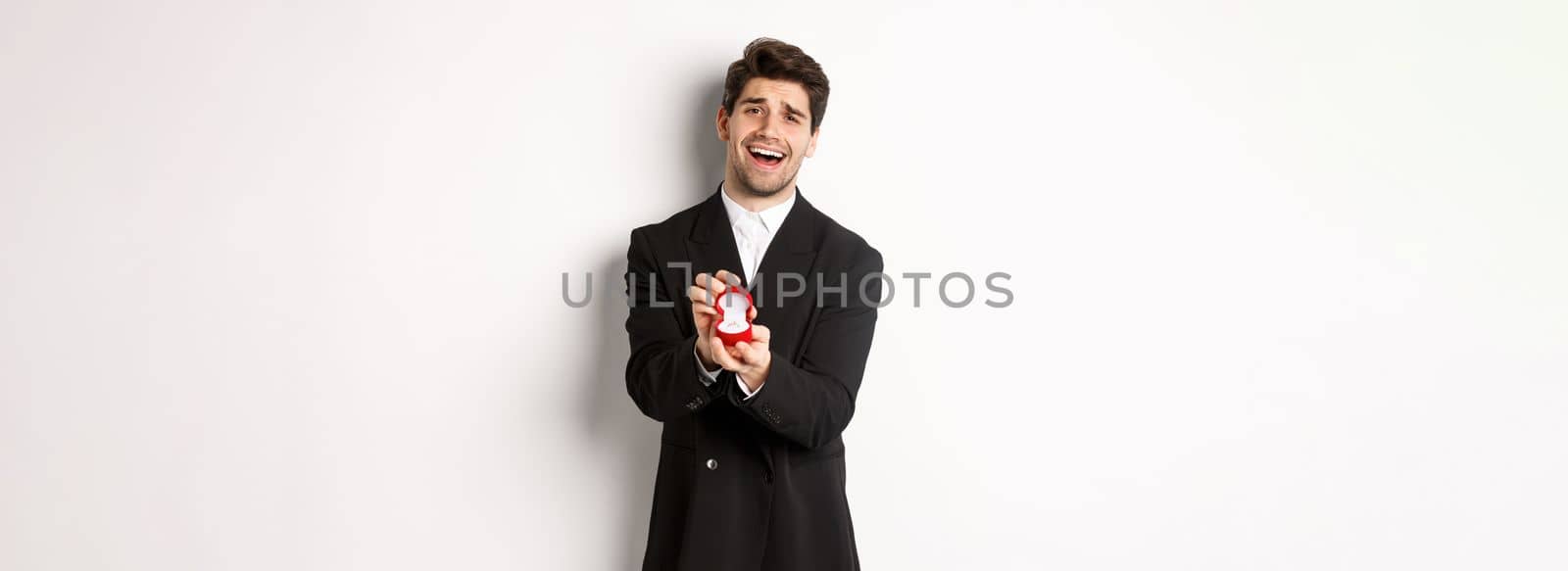 Portrait of handsome man in black suit, open box with wedding ring, making a proposal, asking to marry him, standing against white background.