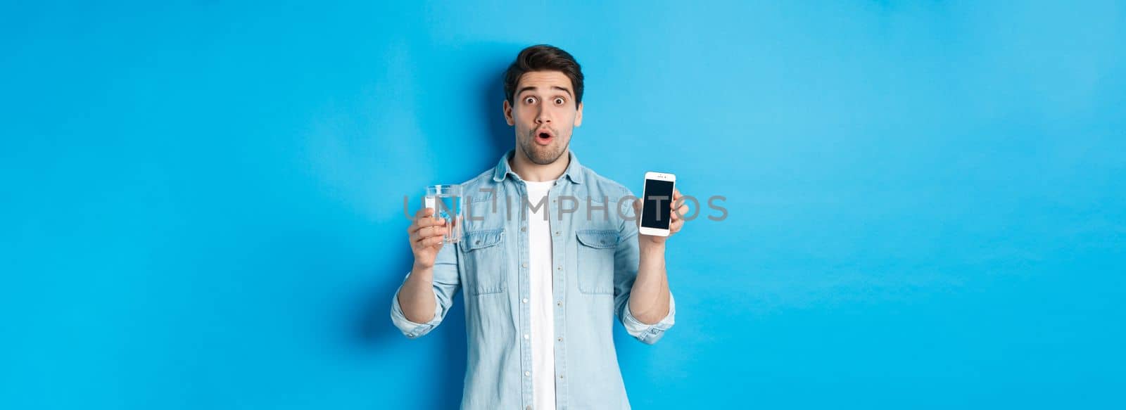 Man looking surprised, showing smartphone screen and glass of water, standing over blue background.