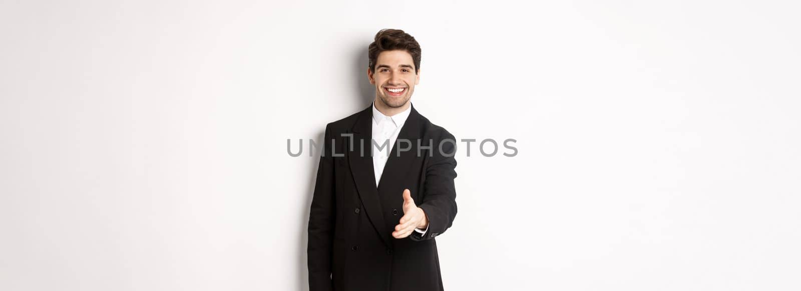 Portrait of handsome businessman in black suit, extending hand for handshake, greet business partners and smiling, welcome to company, standing over white background.