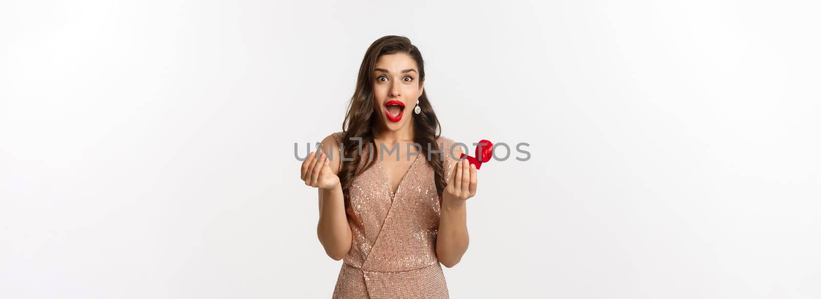 Excited woman holding engagement ring and red box, looking surprised and amazed, receive marriage proposal on date, wearing elegant dress, standing over white background.