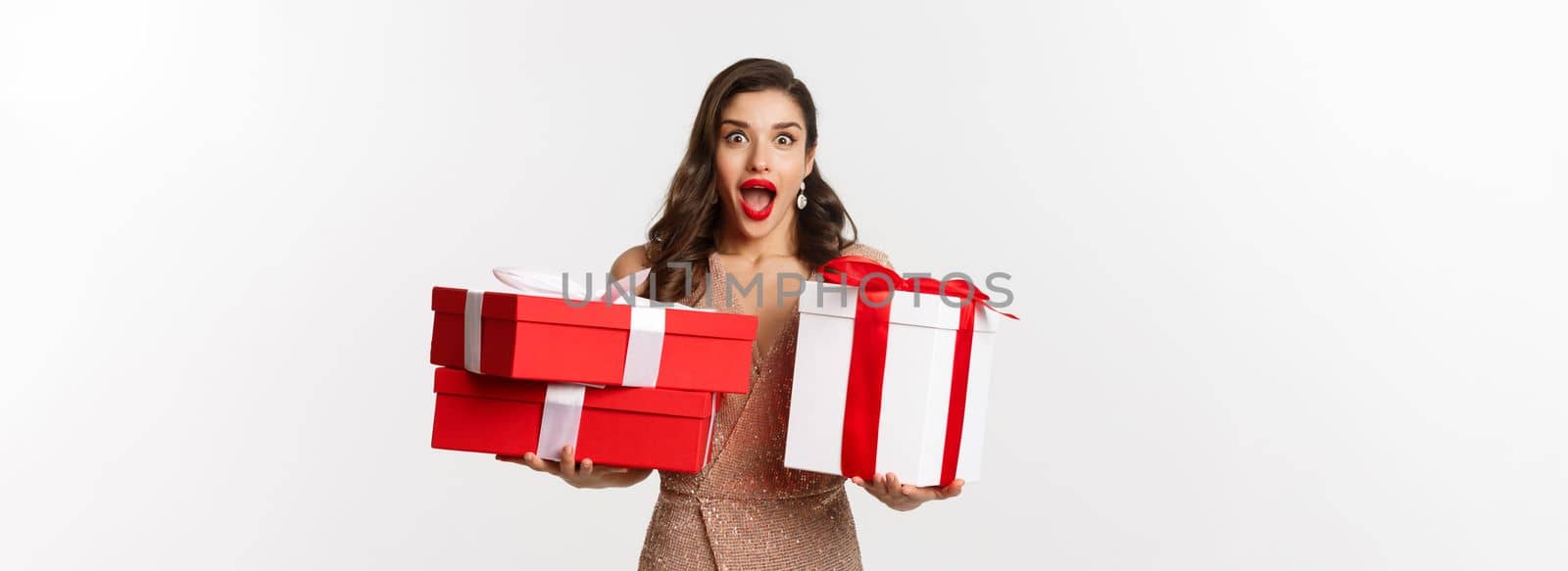 Holidays, celebration concept. Excited and surprised woman holding Christmas gifts and smiling amazed, wearing glamour dress, standing over white background.