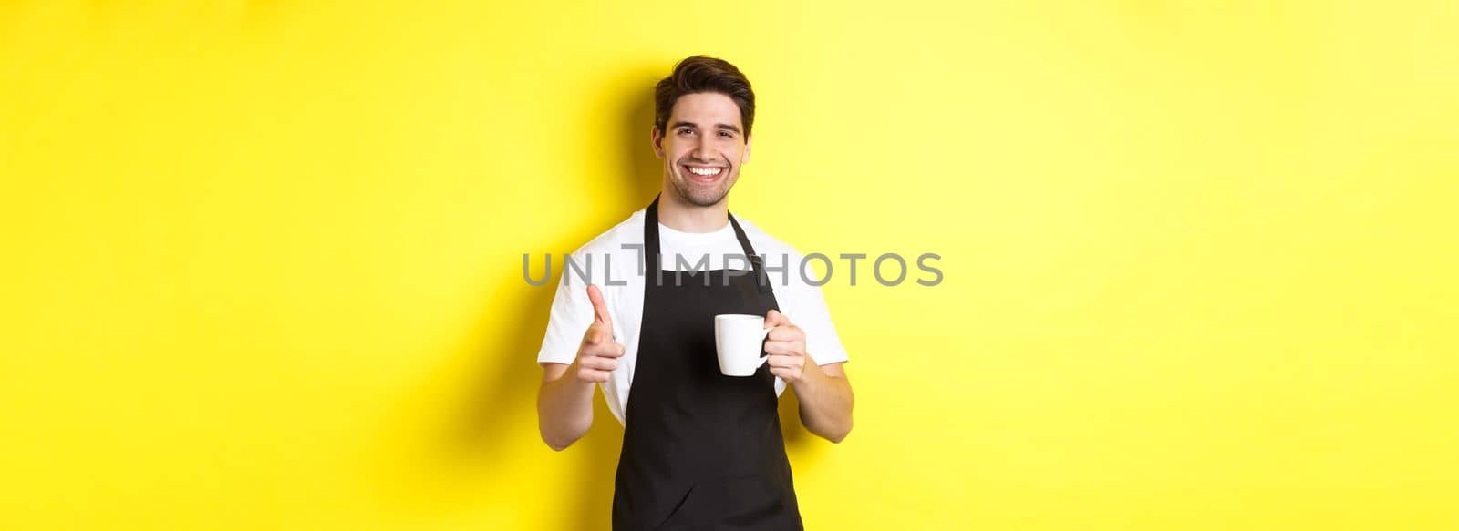 Barista bringing coffee and pointing finger gun at camera, standing in black apron against yellow background.