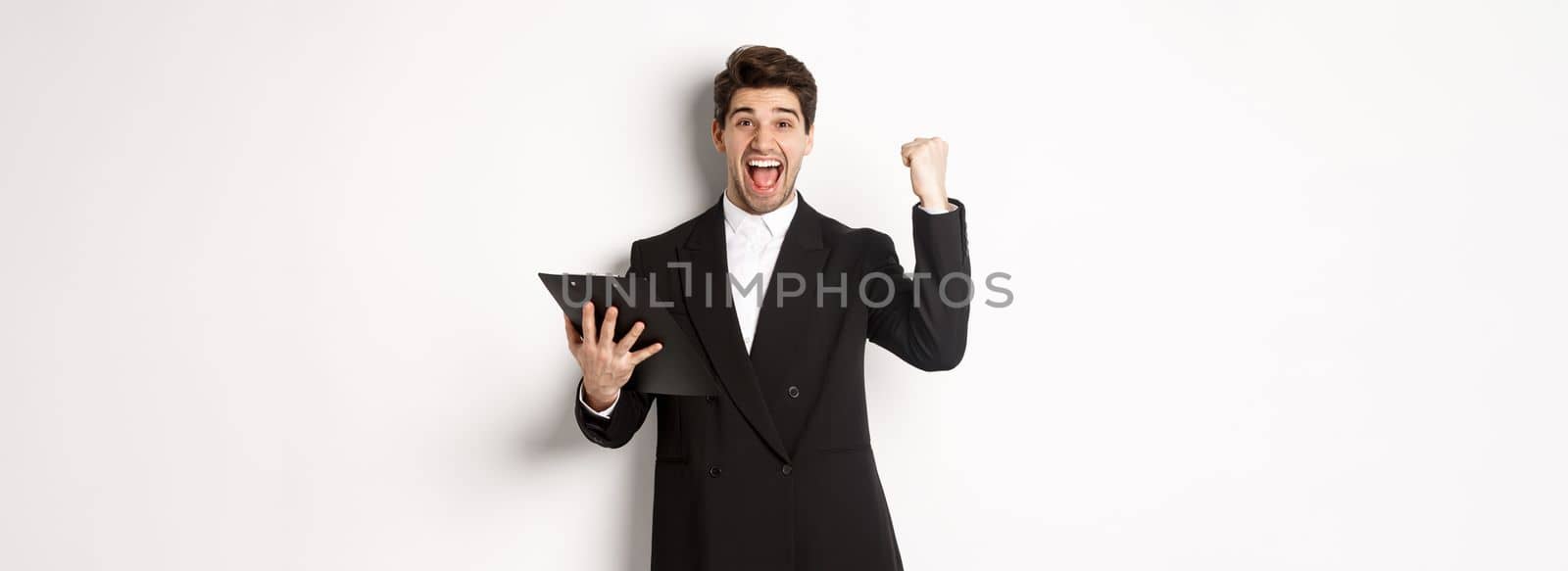 Portrait of excited handsome businessman in black suit, holding clipboard and making fist pump, achieve goal and rejoicing, standing over white background.