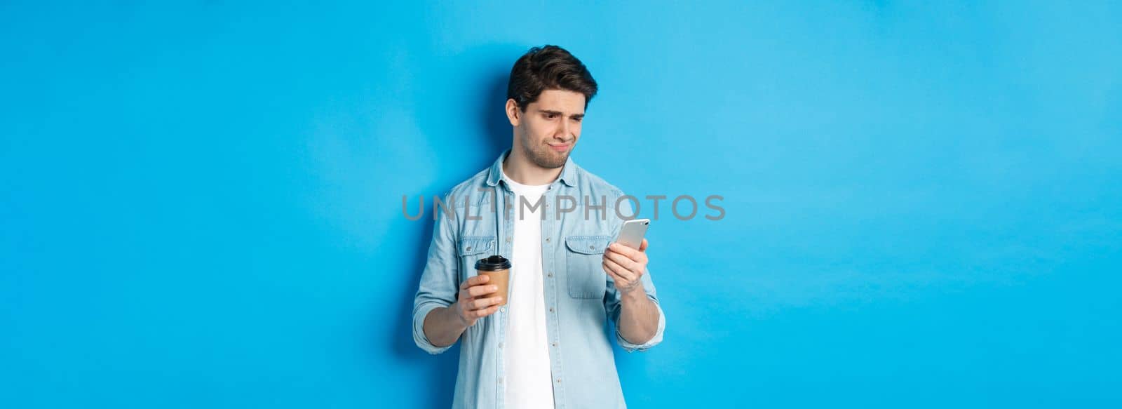 Skeptical and disappointed guy reading upsetting message on phone, holding coffee cup, standing over blue background.