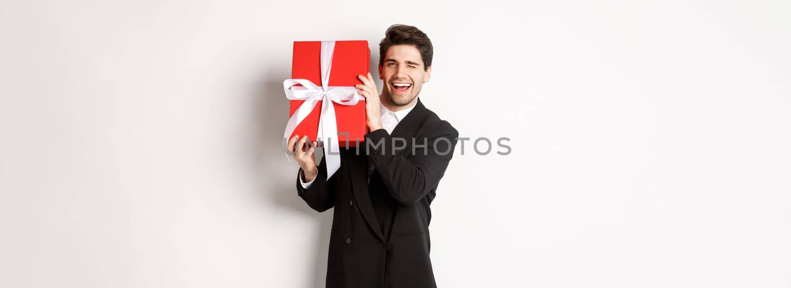 Concept of christmas holidays, celebration and lifestyle. Attractive man in black suit, holding new year gift and smiling, standing with a present over white background.