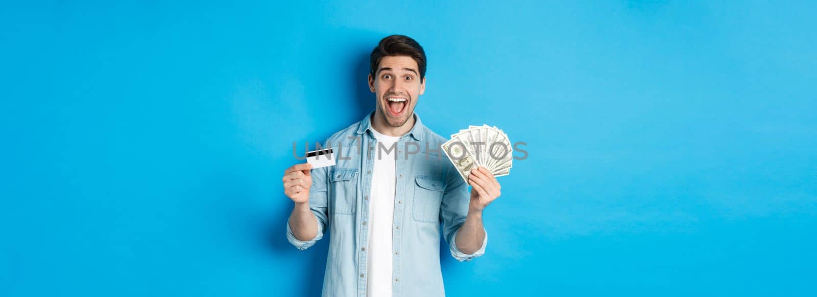 Happy attractive man looking amazed, showing cash and credit card, concept of banks, credit and finance. Blue studio background.