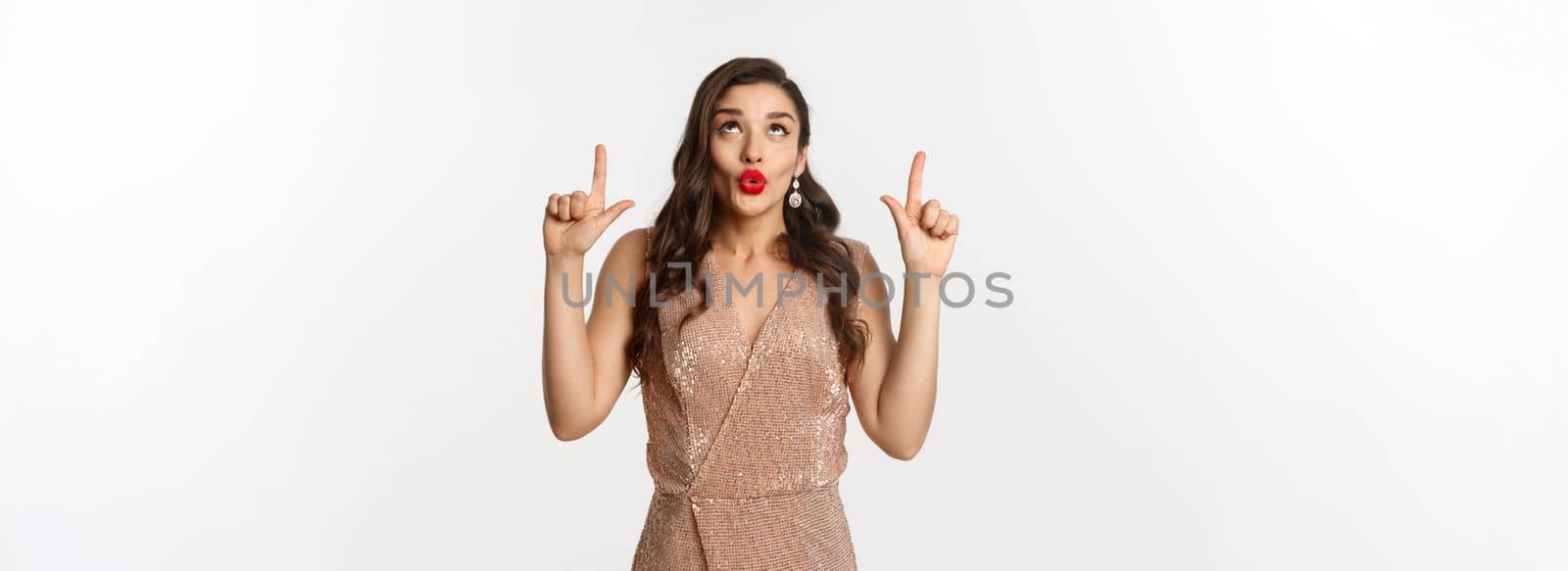 New Year, christmas and celebration concept. Beautiful curious woman in party dress, saying wow and looking at promo offer, pointing fingers up, showing advertisement.