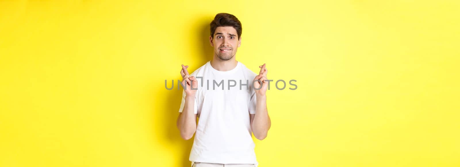 Nervous guy crossing fingers for good luck, hoping for something, standing over yellow background.