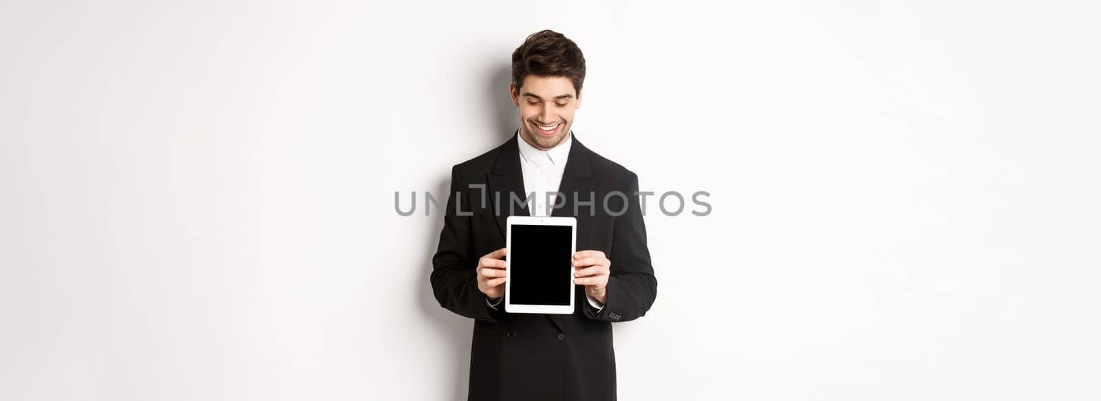 Image of good-looking male entrepreneur in black suit, looking down at digital tablet screen and showing advertisement, standing against white background by Benzoix