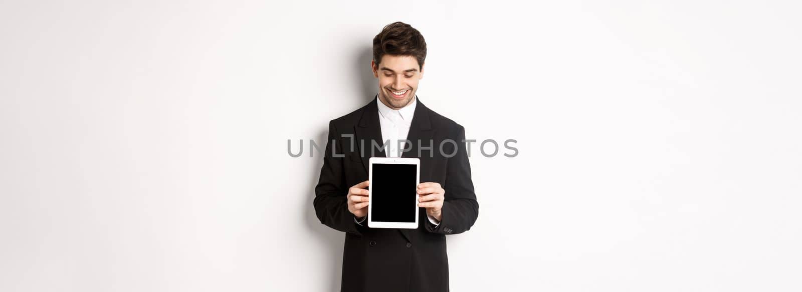 Image of good-looking male entrepreneur in black suit, looking down at digital tablet screen and showing advertisement, standing against white background.