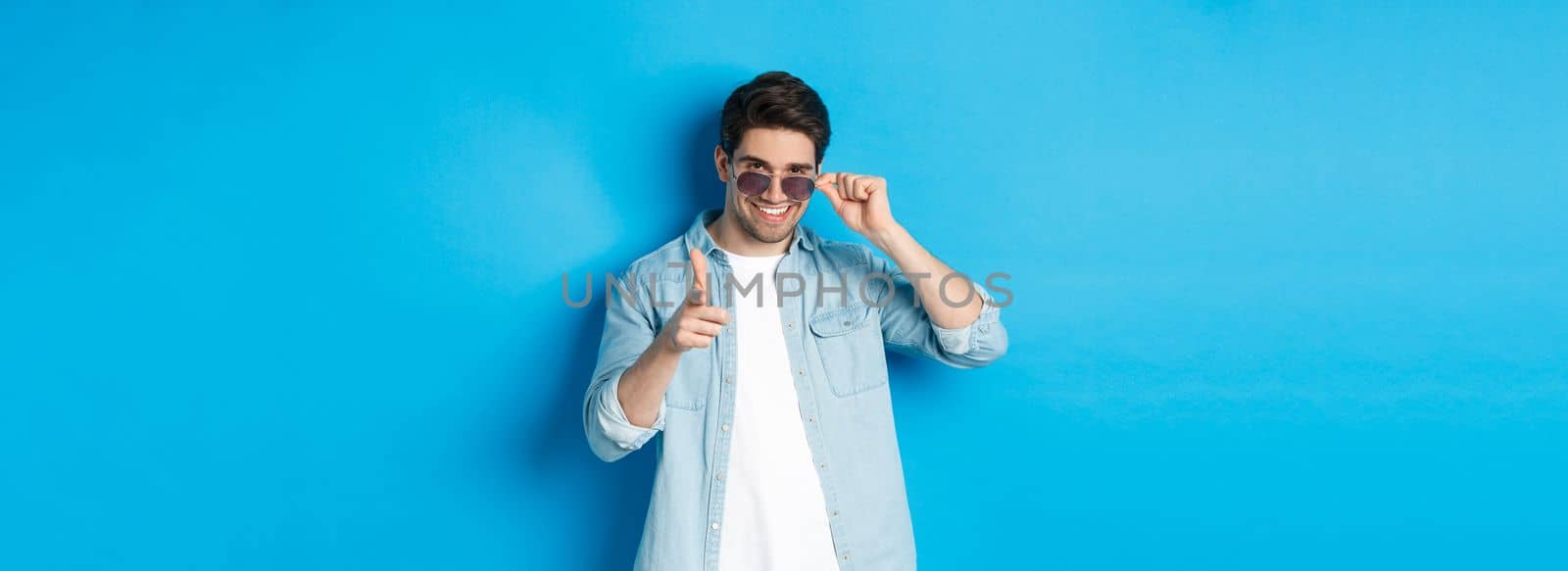 Handsome cheeky guy in sunglasses, smiling and pointing finger gun at camera, flirting with you, standing over blue background.