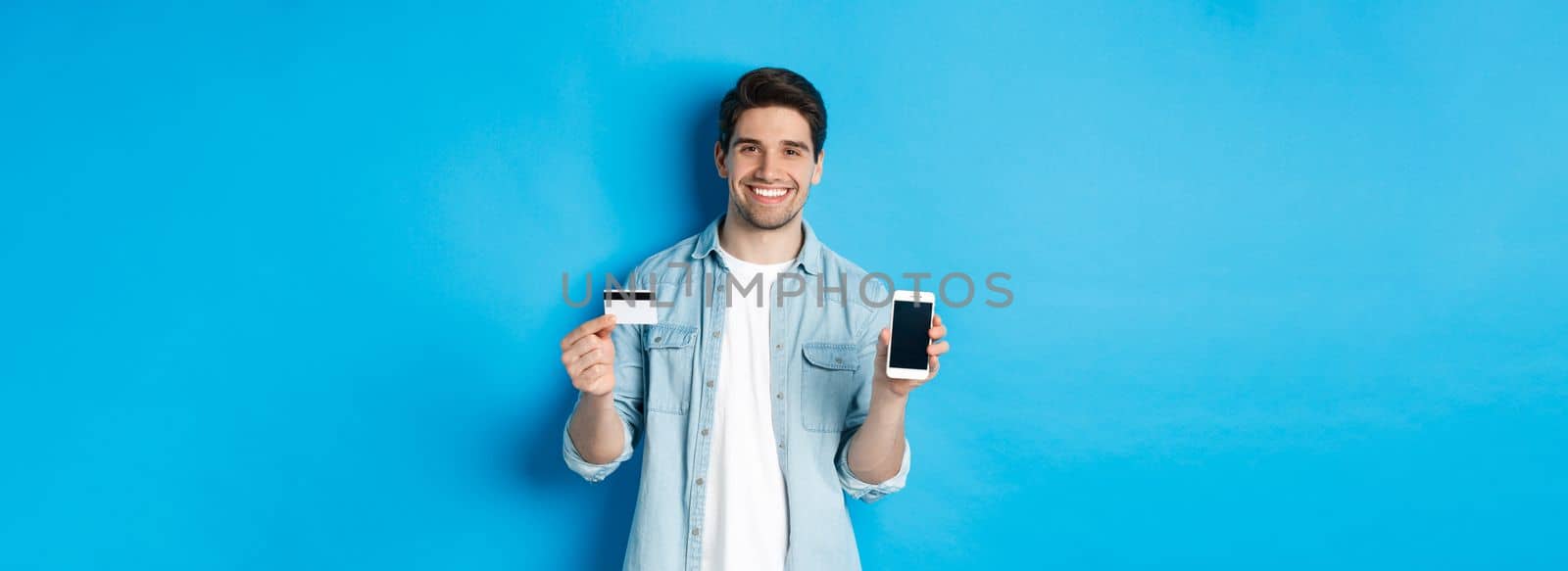 Young smiling man showing smartphone screen and credit card, concept of online shopping or banking.