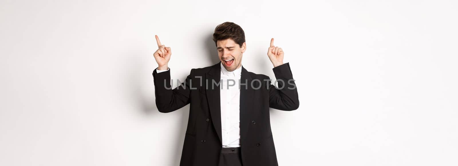 Image of successful handsome businessman dancing, enjoying party, pointing fingers up and having fun, standing over white background, wearing black suit.