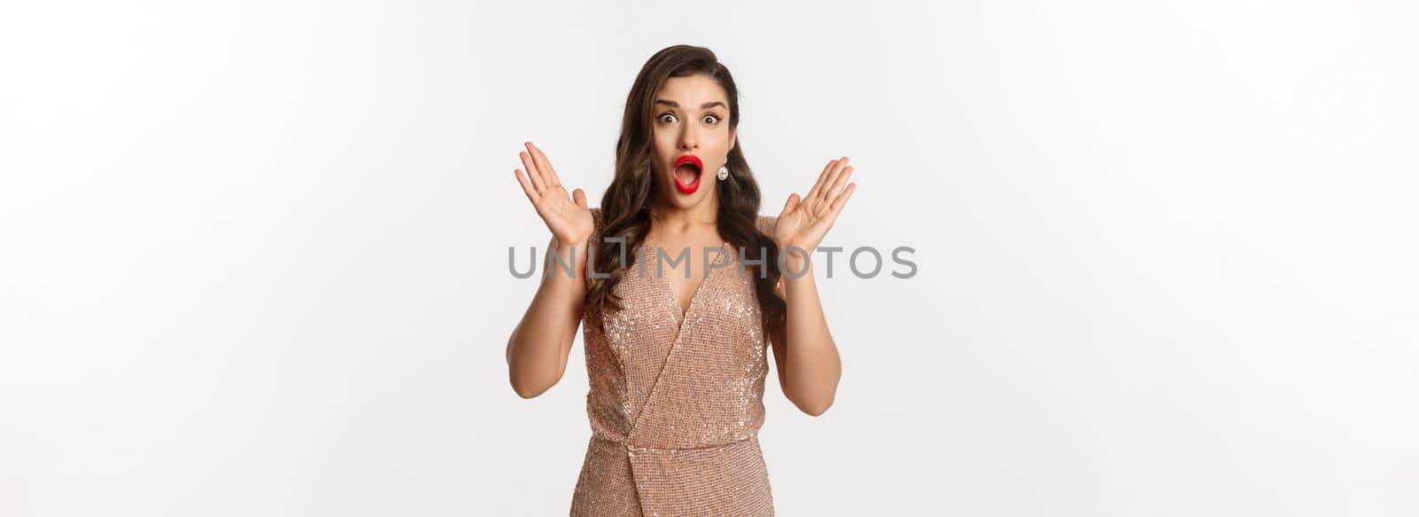 Winter holidays. Beautiful woman in evening dress and makeup, looking surprised and impressed at camera, stare at New Year promo offer, white background.