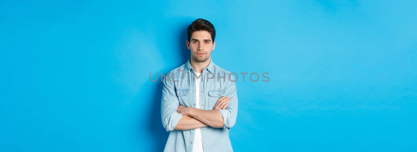 Image of handsome caucasian man in casual outfit, looking serious and confident, standing against blue background.