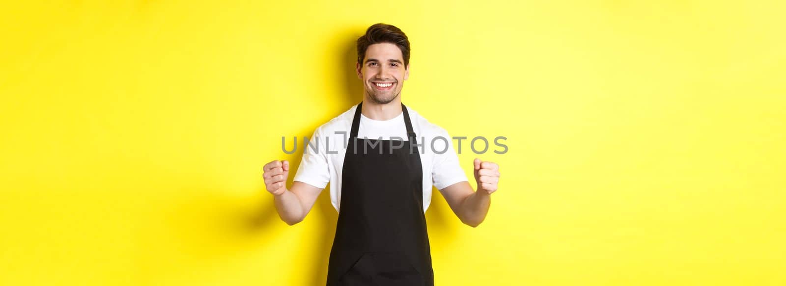 Friendly coffee shop waiter standing with raised hands, place for your sign or logo, standing over yellow background.