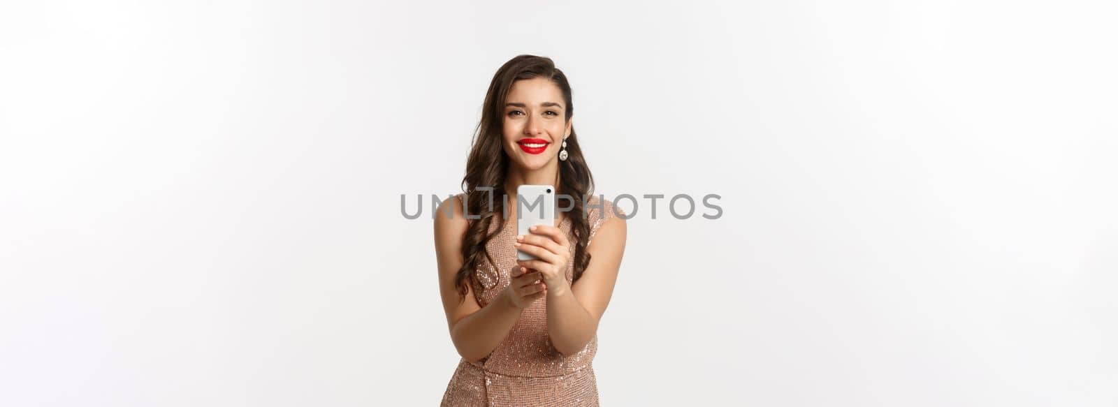 Beautiful young woman in party dress and makeup, taking photo on smartphone, shooting you on phone camera, standing over white background.