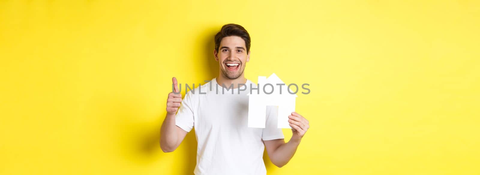 Real estate concept. Happy young man showing paper house model and thumbs up, recommending broker, standing over yellow background.