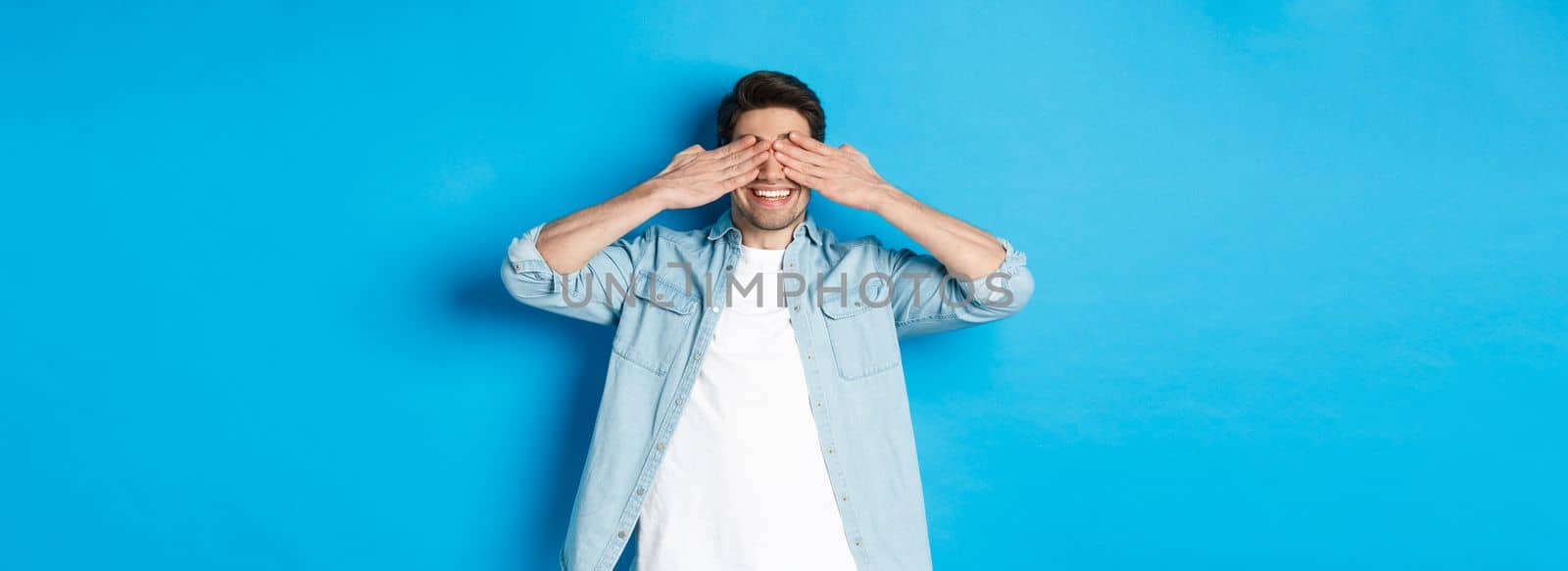 Smiling adult man waiting for surprise, covering eyes with hands and anticipating, standing against blue background in casual clothes.
