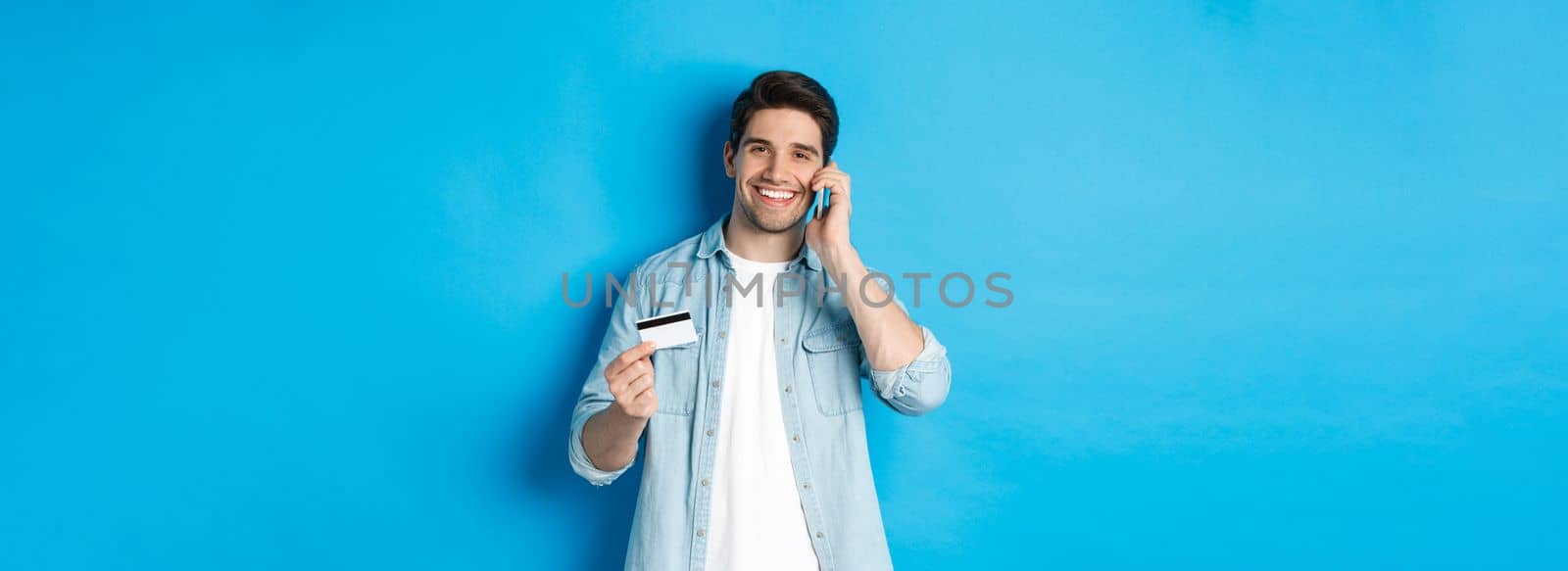 Handsome man calling bank and holding credit card, having mobile conversation, standing over blue background.