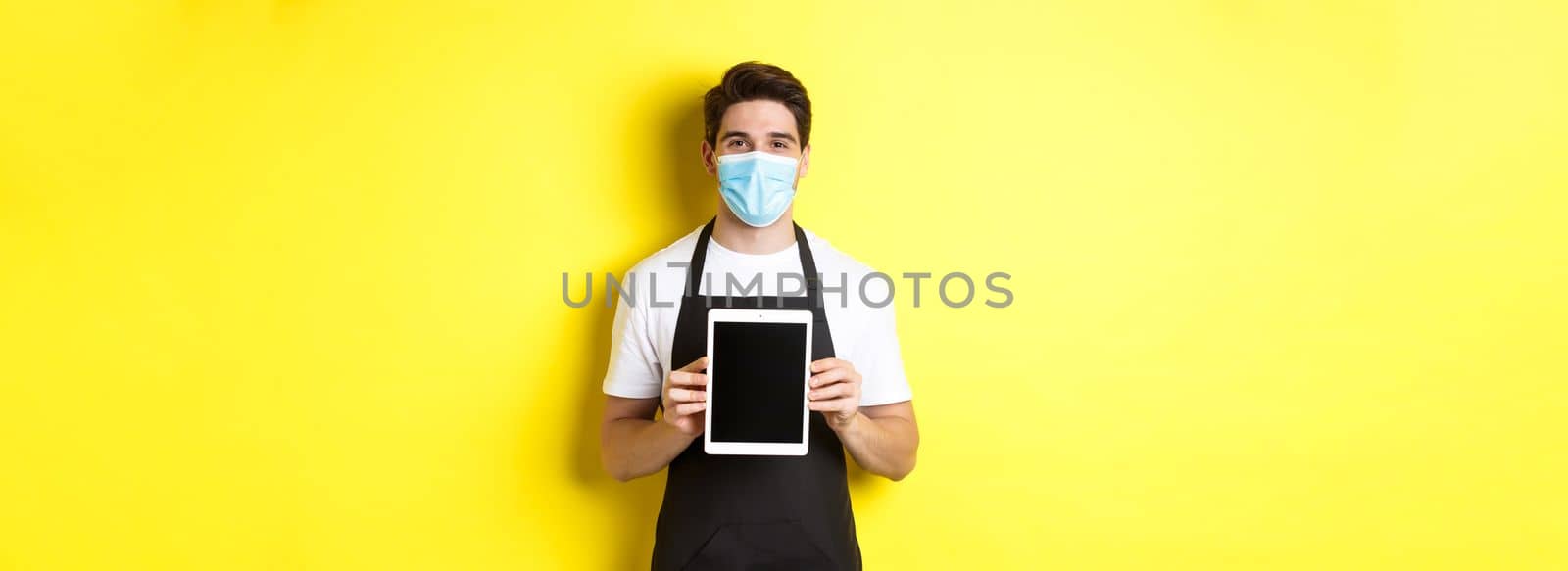 Concept of covid-19, small business and pandemic. Smiling barista in medical mask showing tablet screen, standing over yellow background.