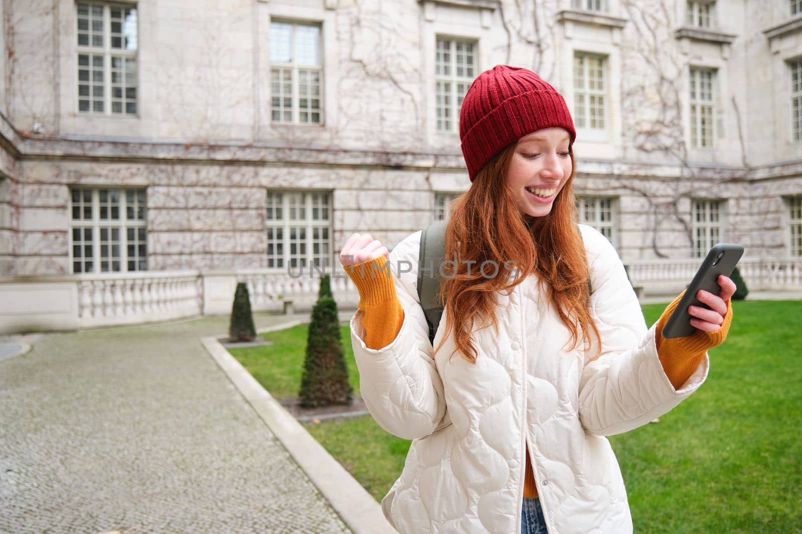 Cheerful redhead girl winning, celebrating victory, read great news on mobile phone and jumping from joy, saying yes, standing outside building in yard.