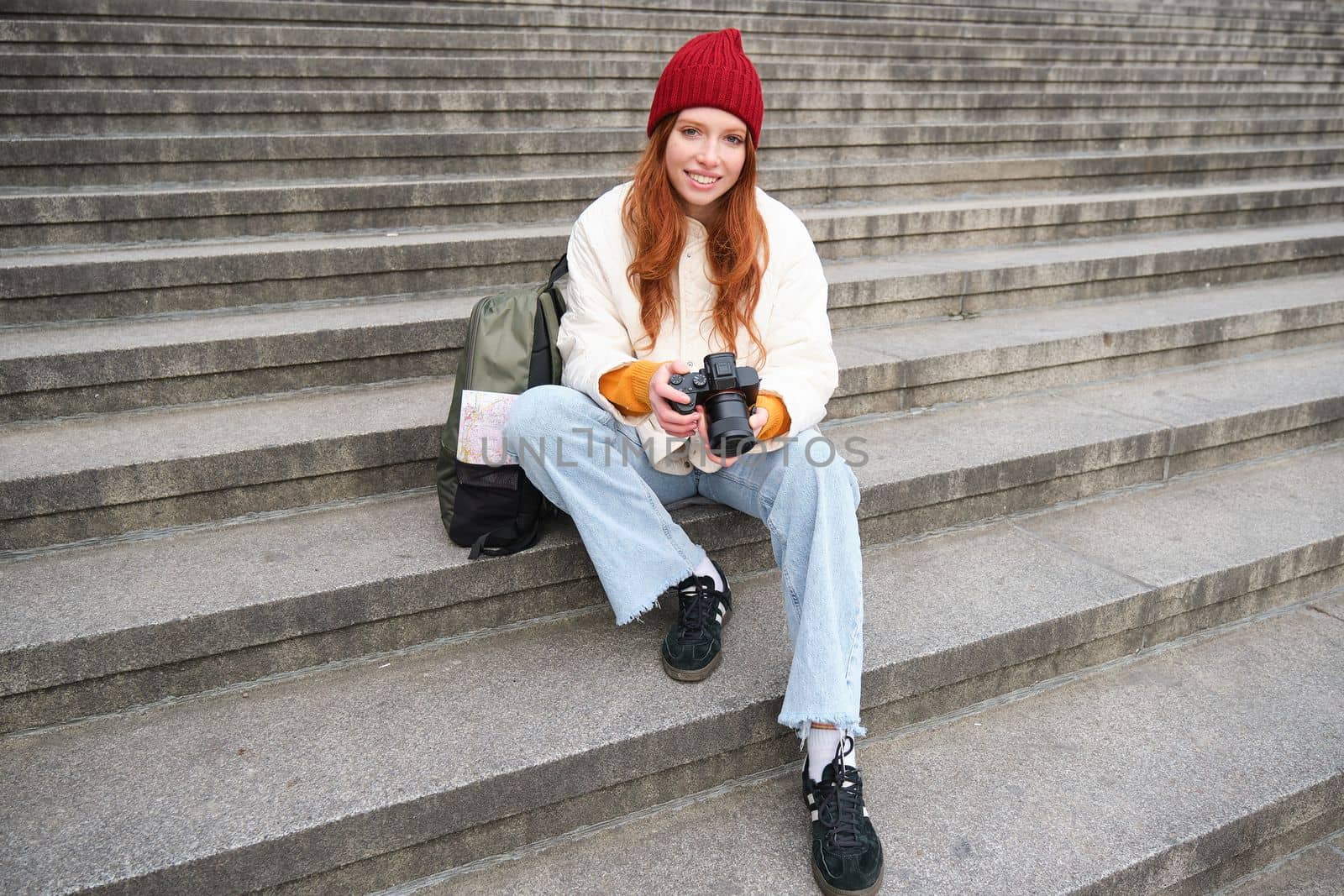 Young student, photographer sits on street stairs and checks her shots on professional camera, taking photos outdoors.