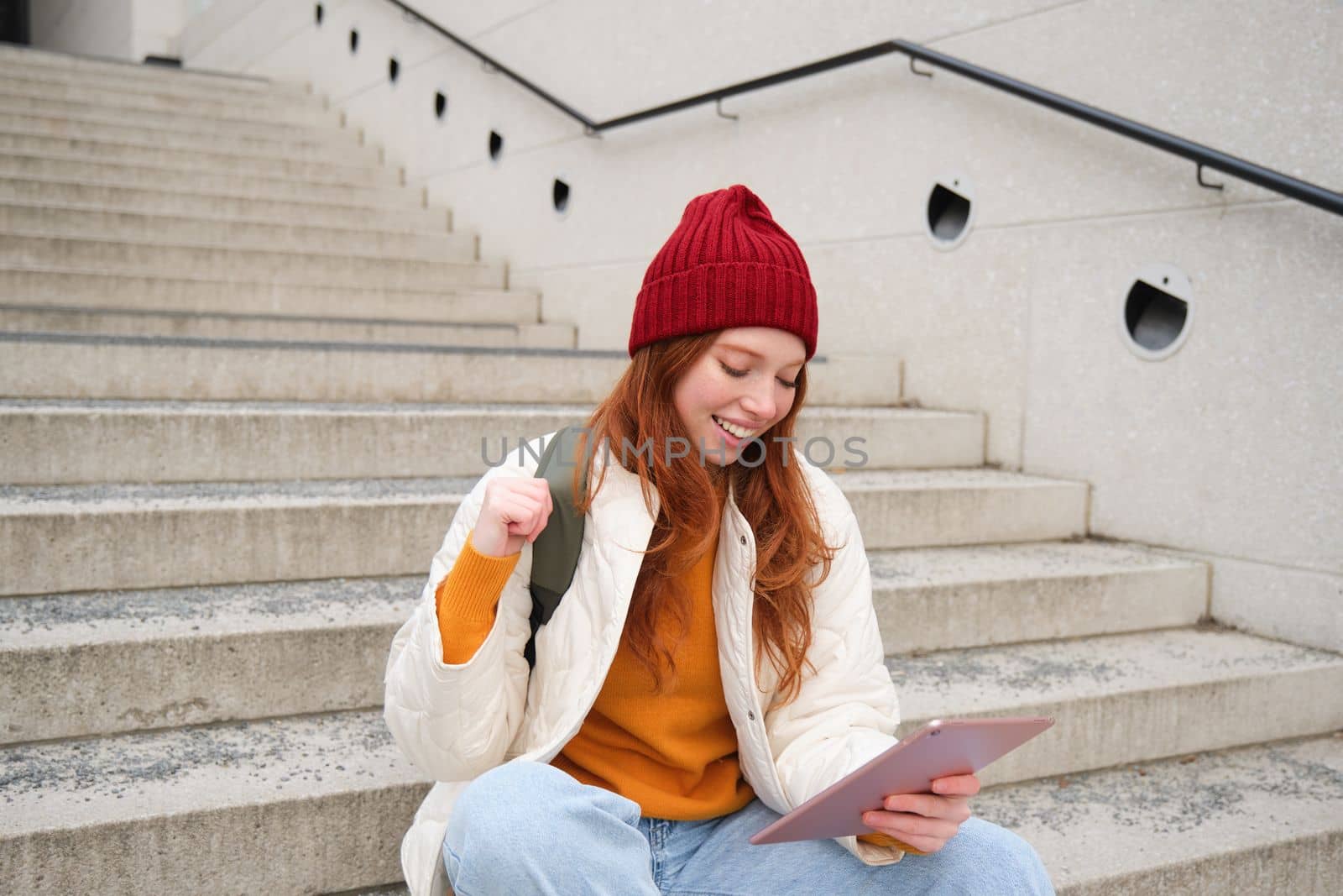 Young beautiful girl with long red hair, smiles, uses internet application, connect to wifi near campus, sits on stairs with digital tablet.