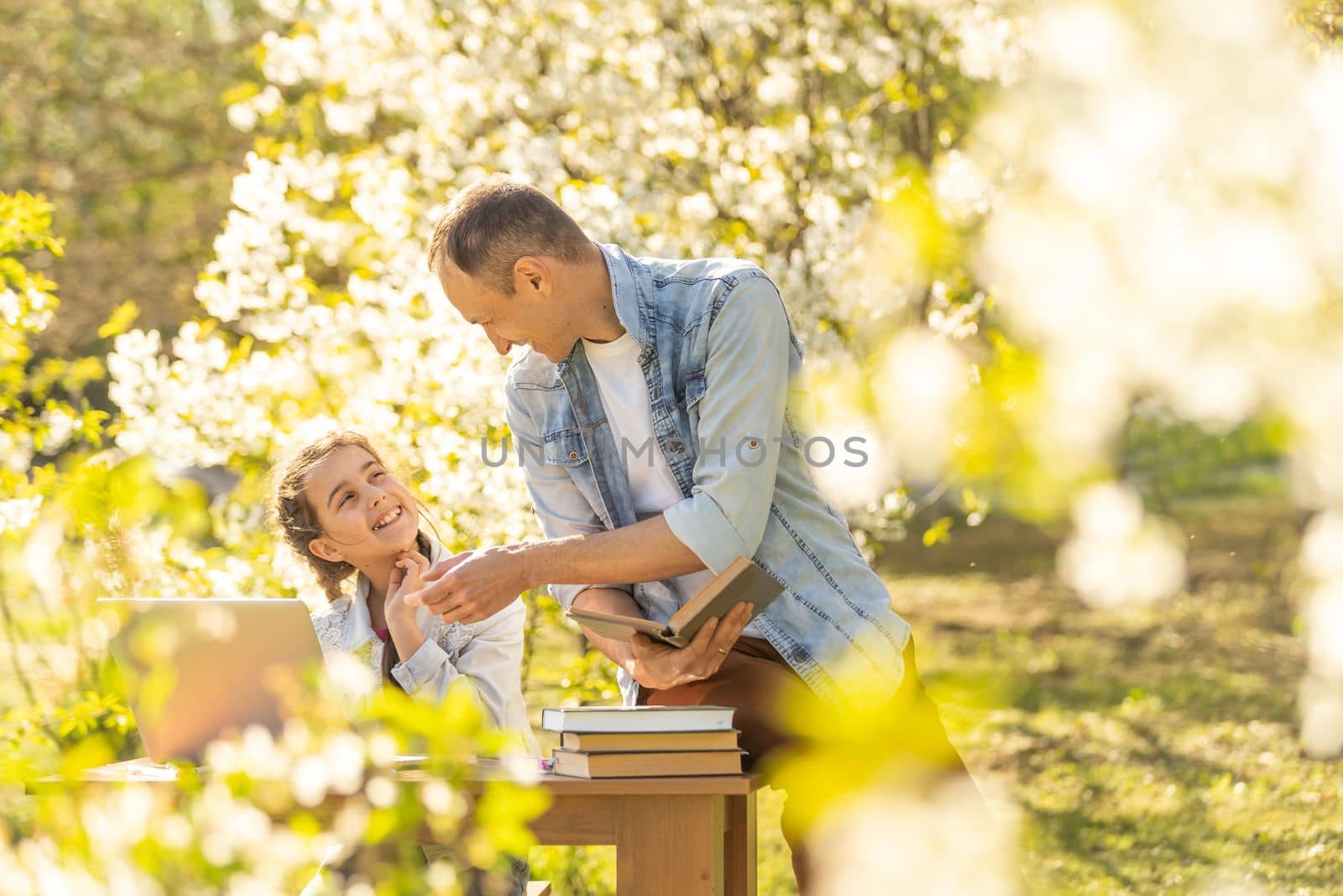A girl and a young father are sitting at a laptop and studying in a flowered garden. Against the background of green grass and flowering trees. Remotely buy items in a store.