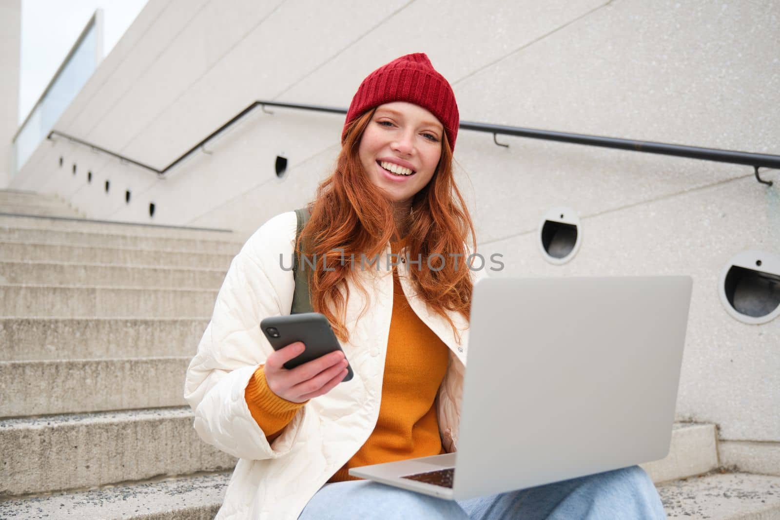 Smiling redhead woman with mobile phone and laptop, sitting on stairs outside building, connects to public wifi, using smartphone and computer.