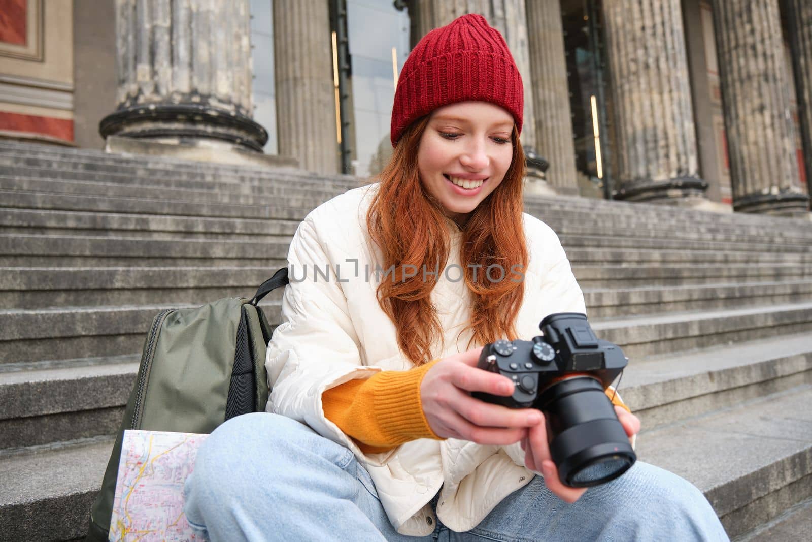 Young student, photographer sits on street stairs and checks her shots on professional camera, taking photos outdoors.