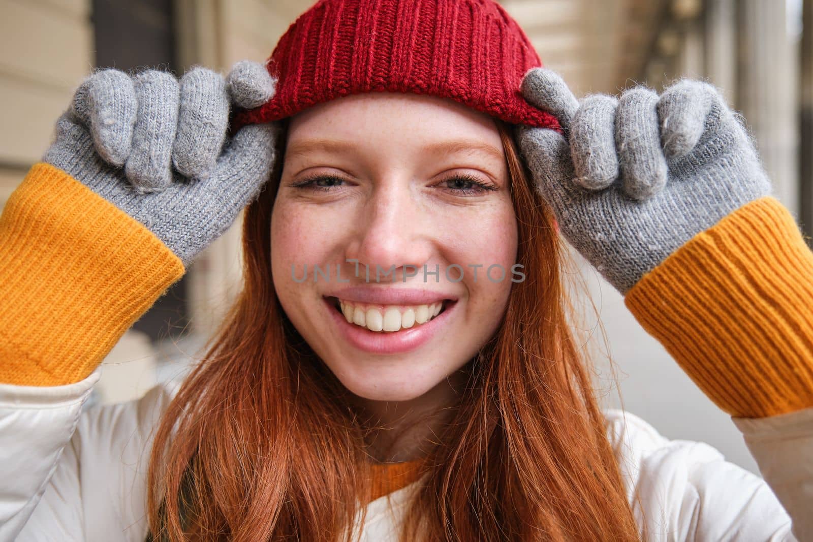 Headshot of happy redhead girl with freckles, wears red hat and gloves in winter, walks around city on chilly weather and smiles.