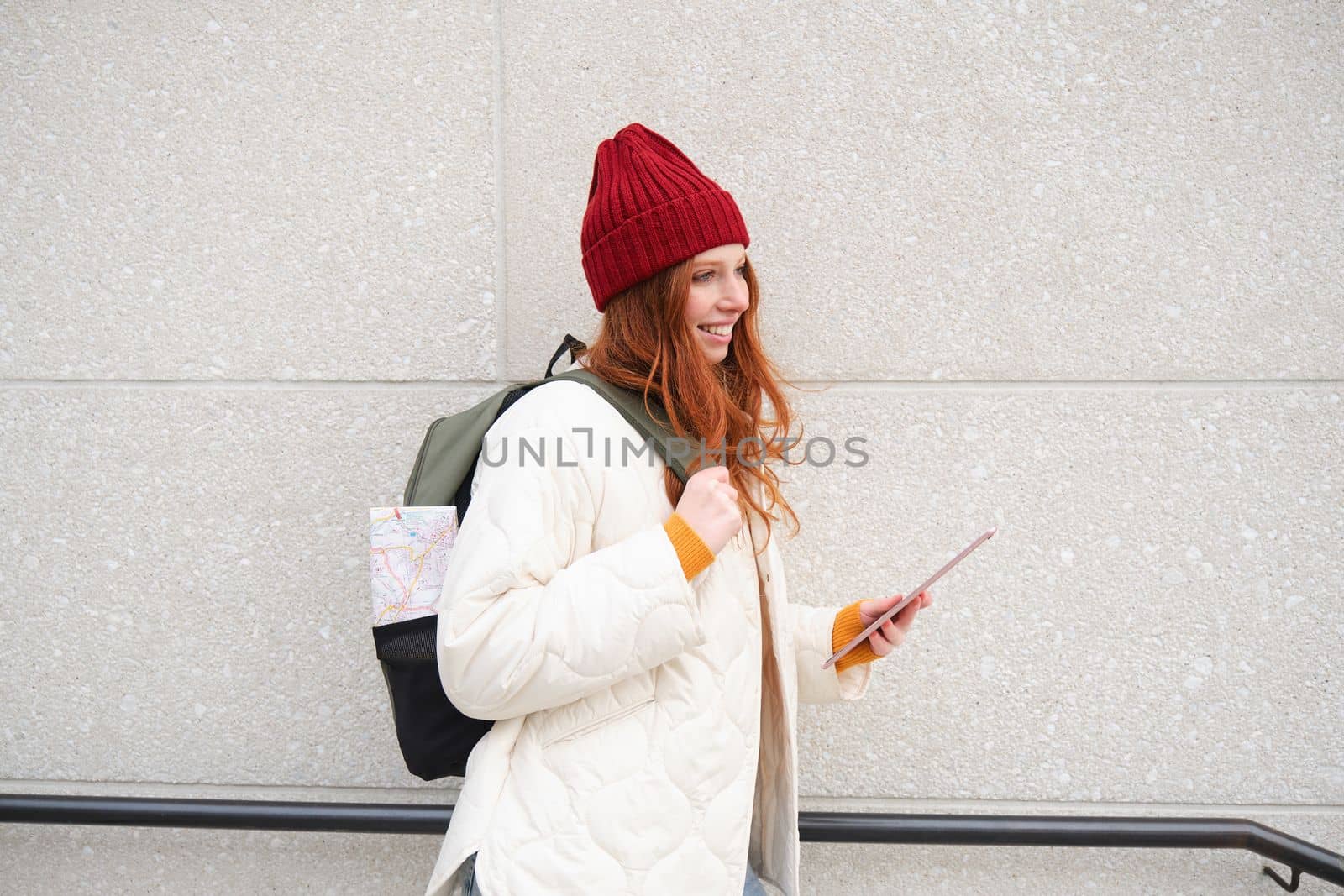Young redhead girl, walks with digital tablet around city, stands on street with backpack and gadget, looks at online map, searches for a place in internet.