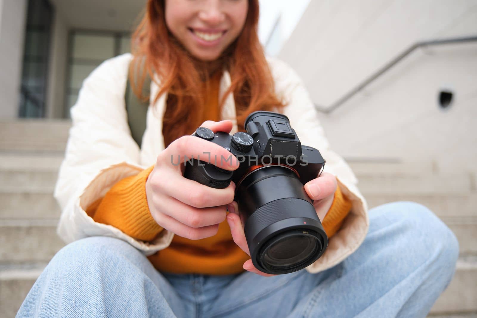 Smiling redhead girl photographer, checks her shots, holds camera and looks at screen, takes photos outdoors, walks around street and does streetstyle shooting.