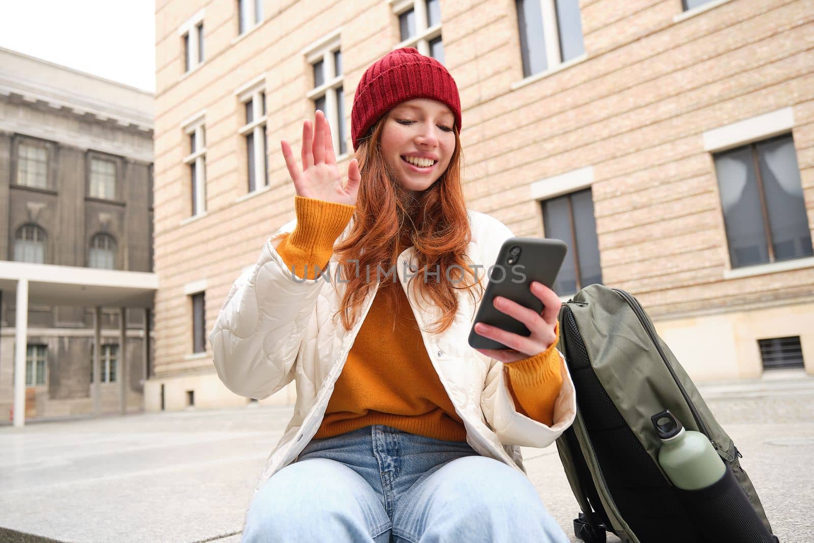 Social media and people. Young redhead girl sits on street, uses mobile phone app, looks up information in internet, holds smartphone.