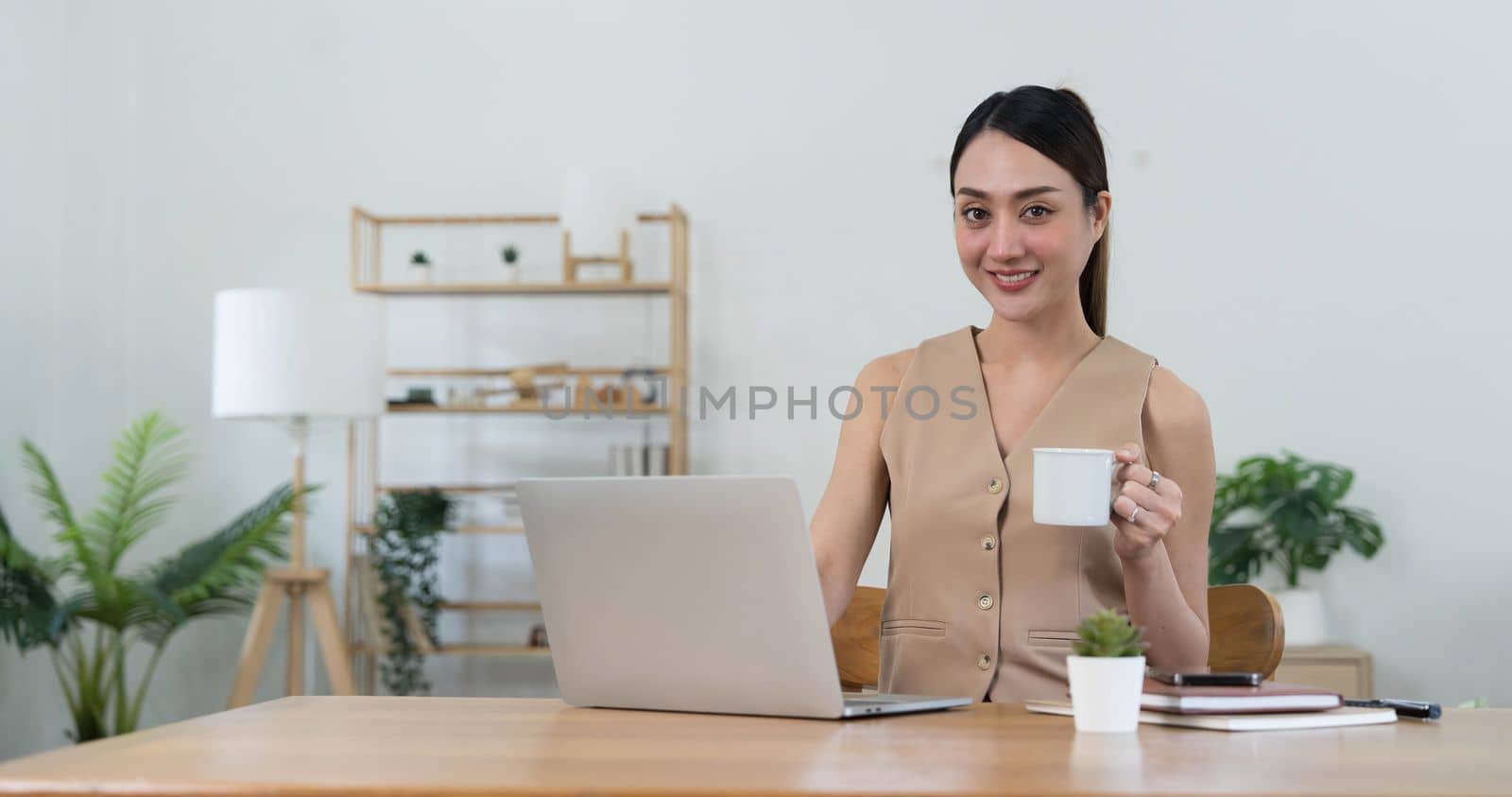 Beautiful young Asian businesswoman smiling holding a coffee mug and laptop working at the office...