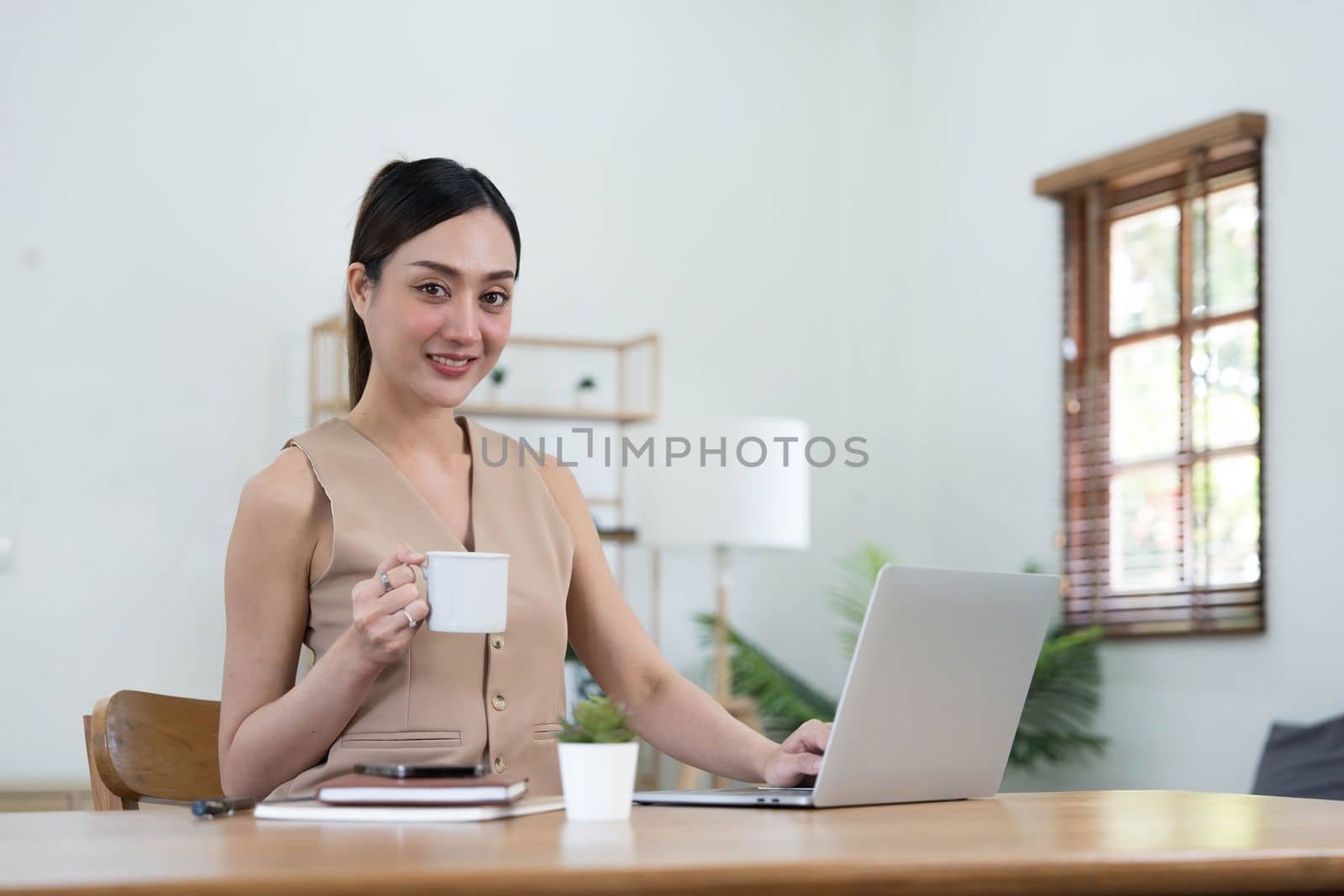 Beautiful young Asian businesswoman smiling holding a coffee mug and laptop working at the office. by wichayada