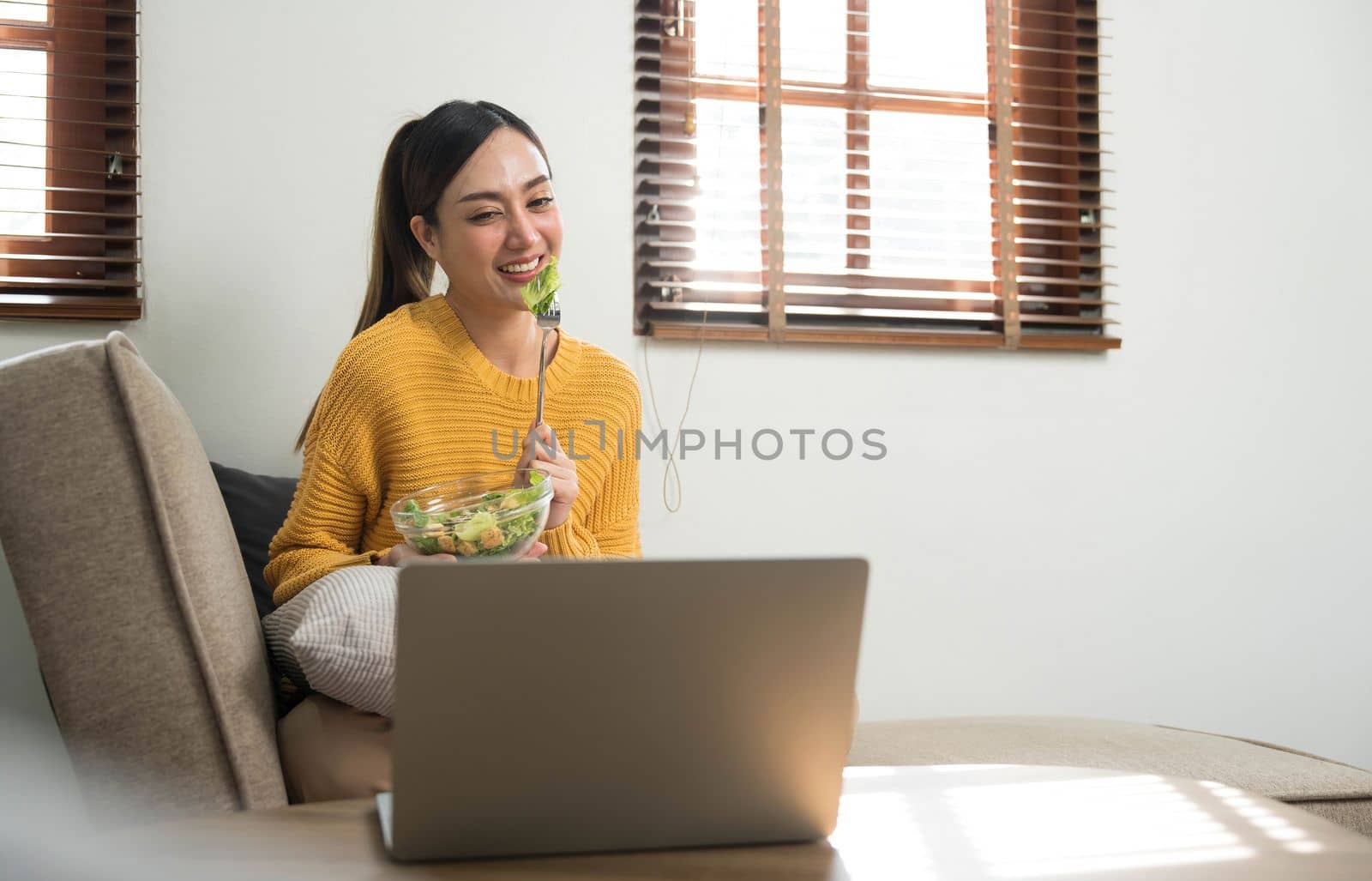 People relax at home and wellness lifestyle. Young adult asian woman eating salad and using laptop computer for watching online movie on internet. by wichayada