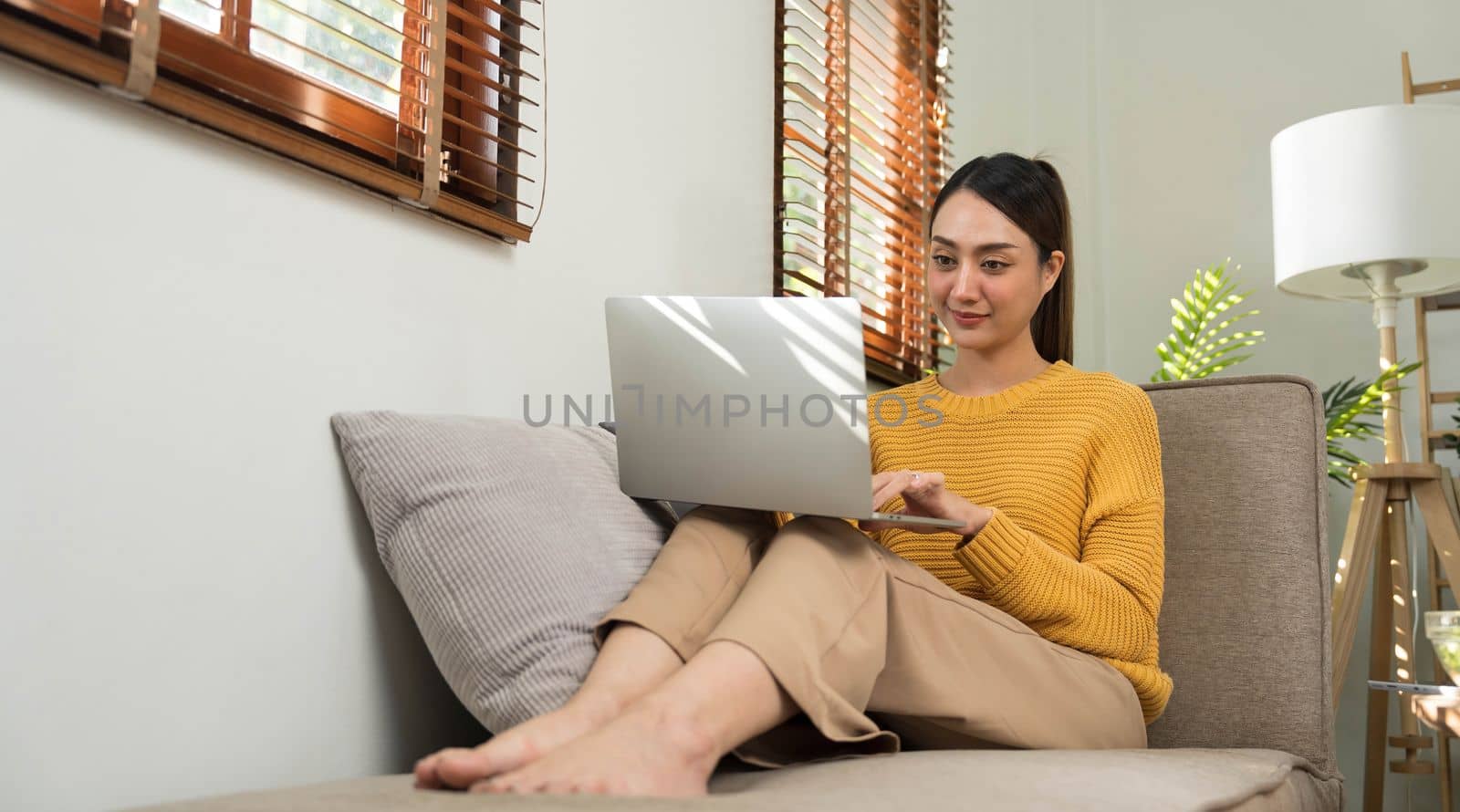 Portrait of a young happy female sitting on the couch and working on project, watching movie on laptop, studying, blogging, resting and chatting online. by wichayada