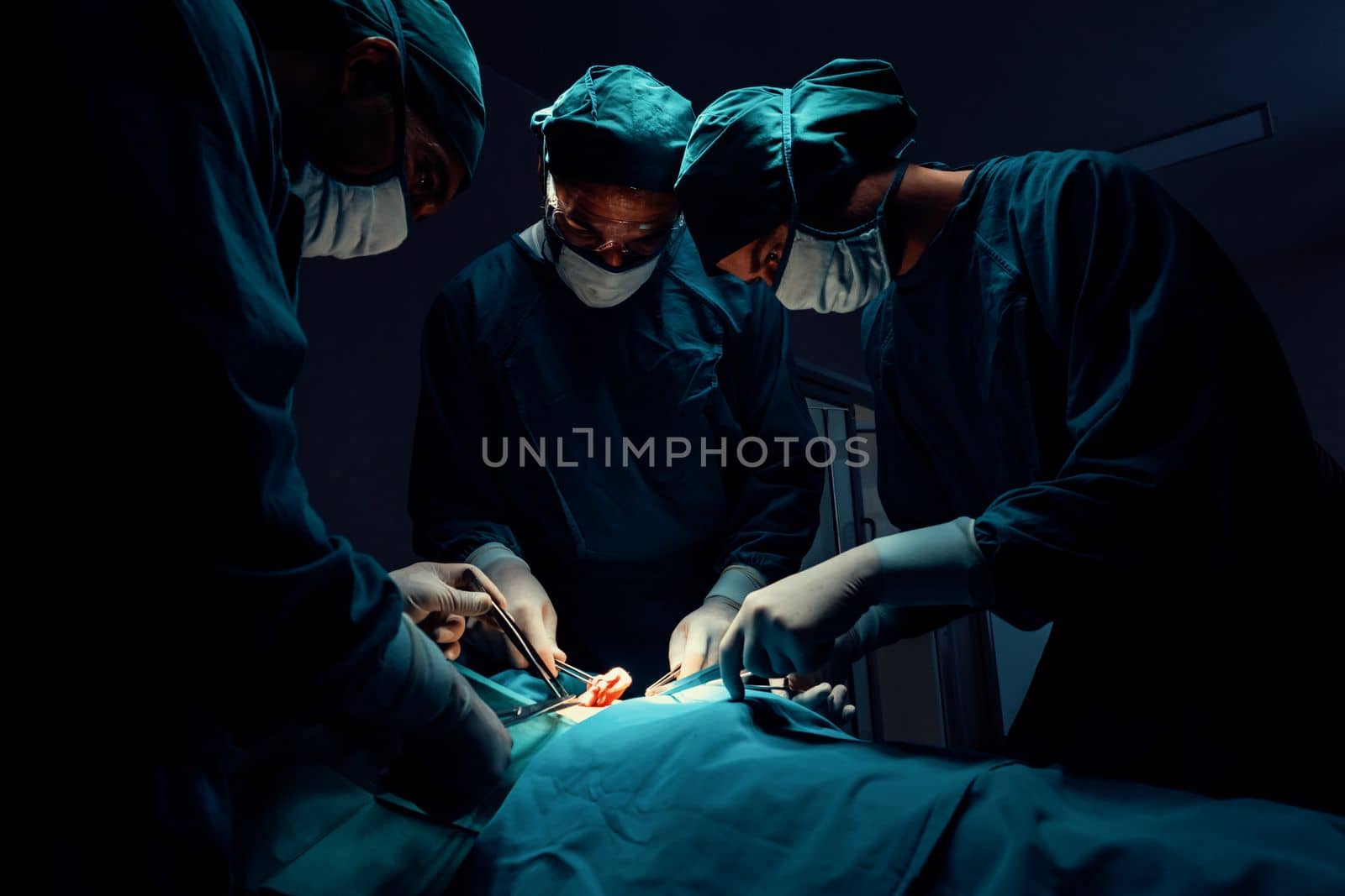 Surgical team performing surgery to patient in sterile operating room. In a surgery room lit by a lamp, a professional and confident surgical team provides medical care to an unconscious patient.