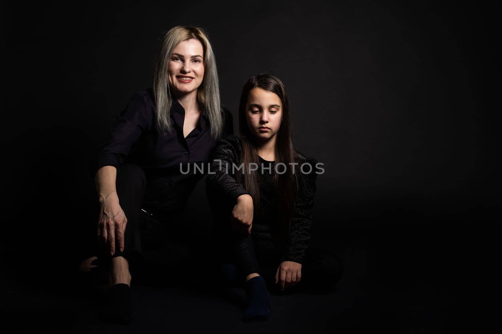 Young mother and her little daughter are looking at camera and smiling, sitting against black wall.
