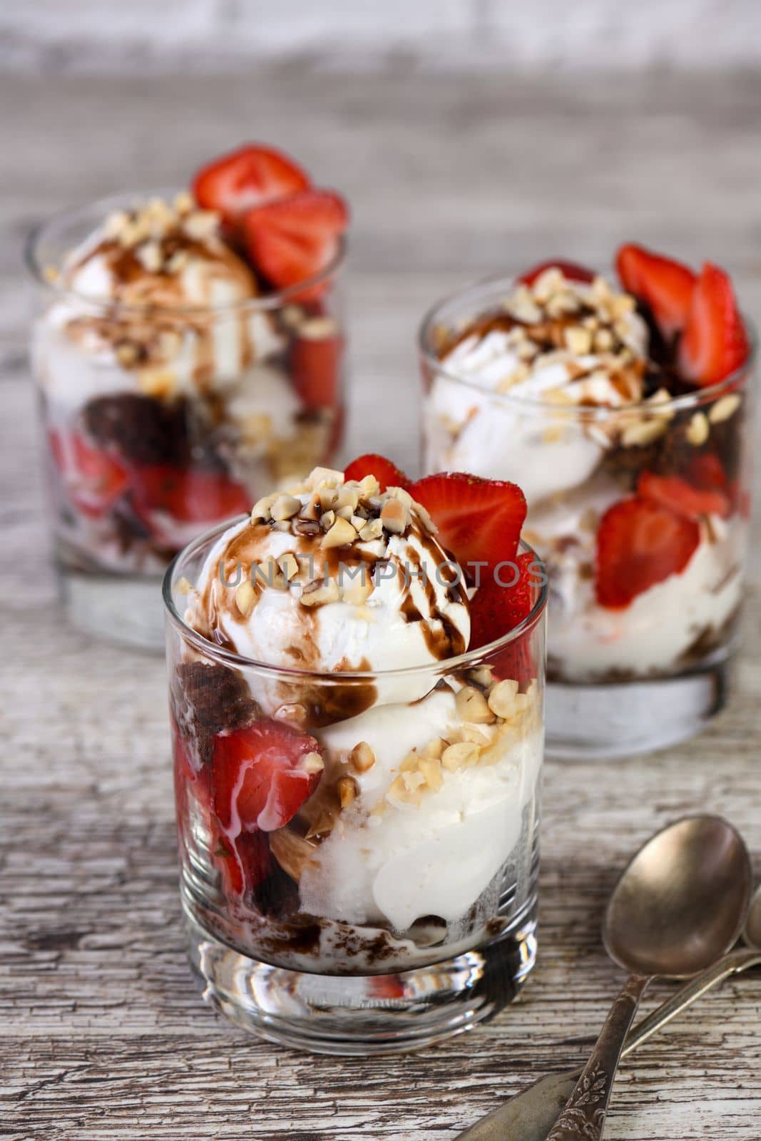 Summer cold dessert - Plombir ice cream scoop with chocolate sauce, cookie crumbs, ripe strawberries, crushed peanut nuts in a glass on a wooden table