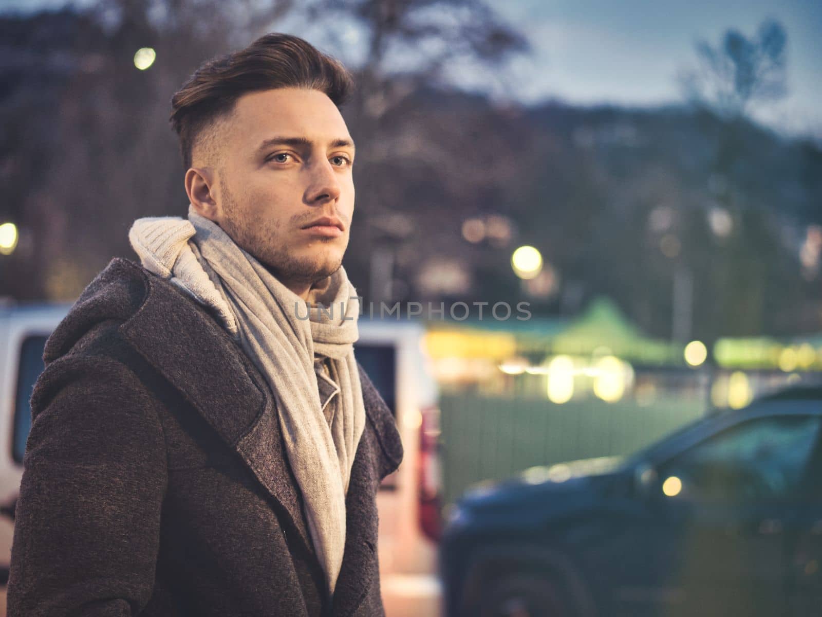 Handsome trendy young man, standing on a sidewalk in city setting at night wearing a fashionable winter coat and scarf, looking at camera