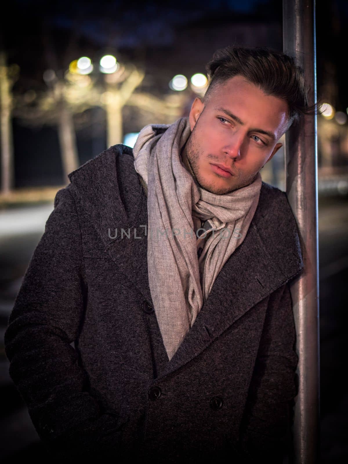 Handsome trendy young man, standing on a sidewalk in city setting at night wearing a fashionable winter coat and scarf