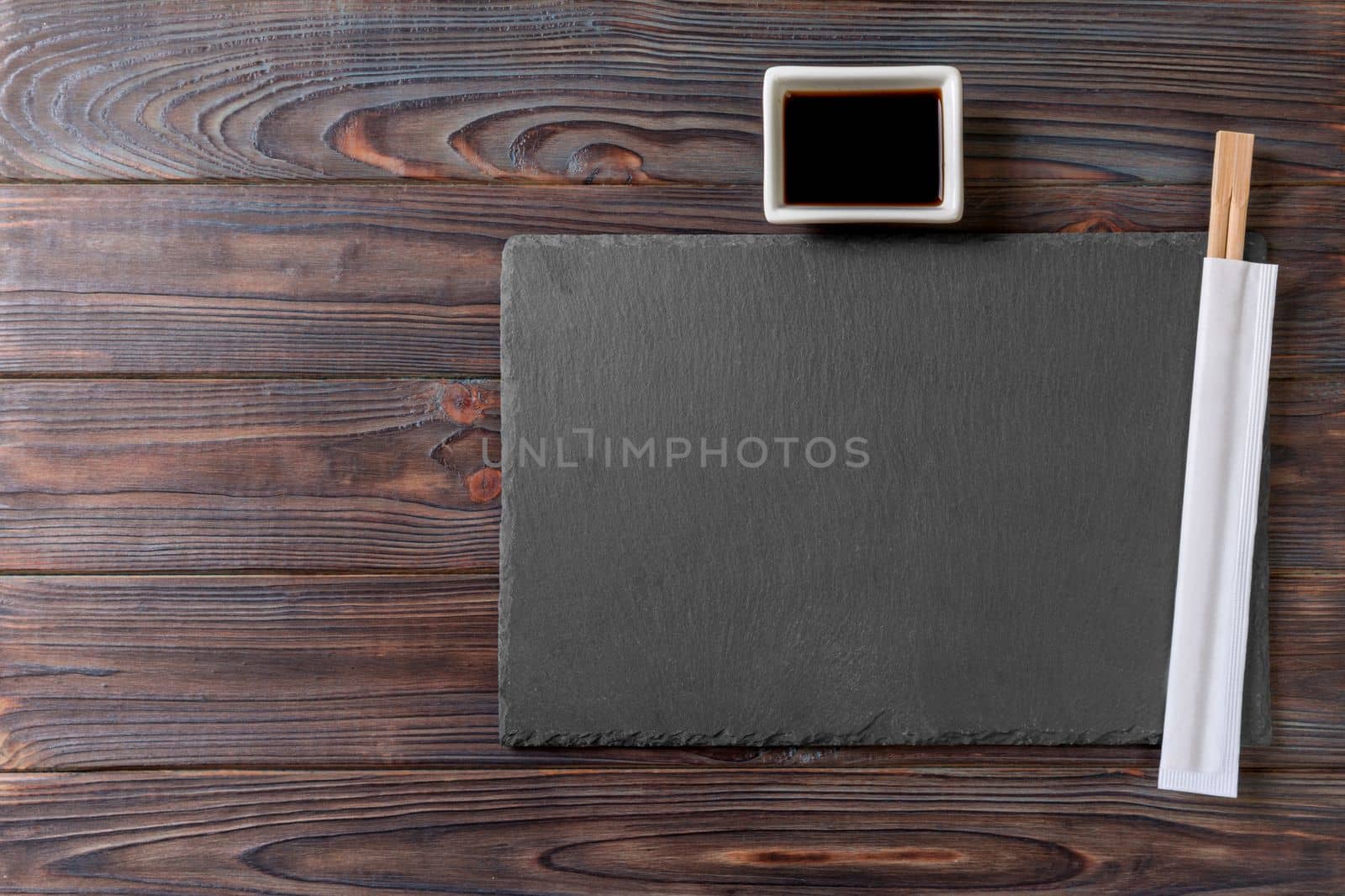 Empty rectangular black slate plate with chopsticks for sushi and soy sauce on wooden background. Top view with copy space.