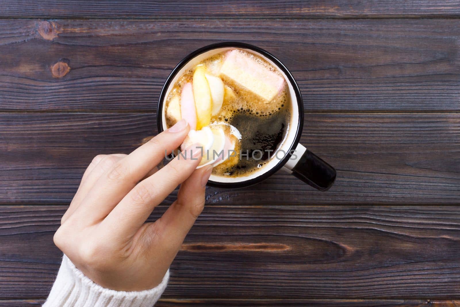 female hand holding cup of hot cocoa or chocolate with marshmallow on wooden table from above.