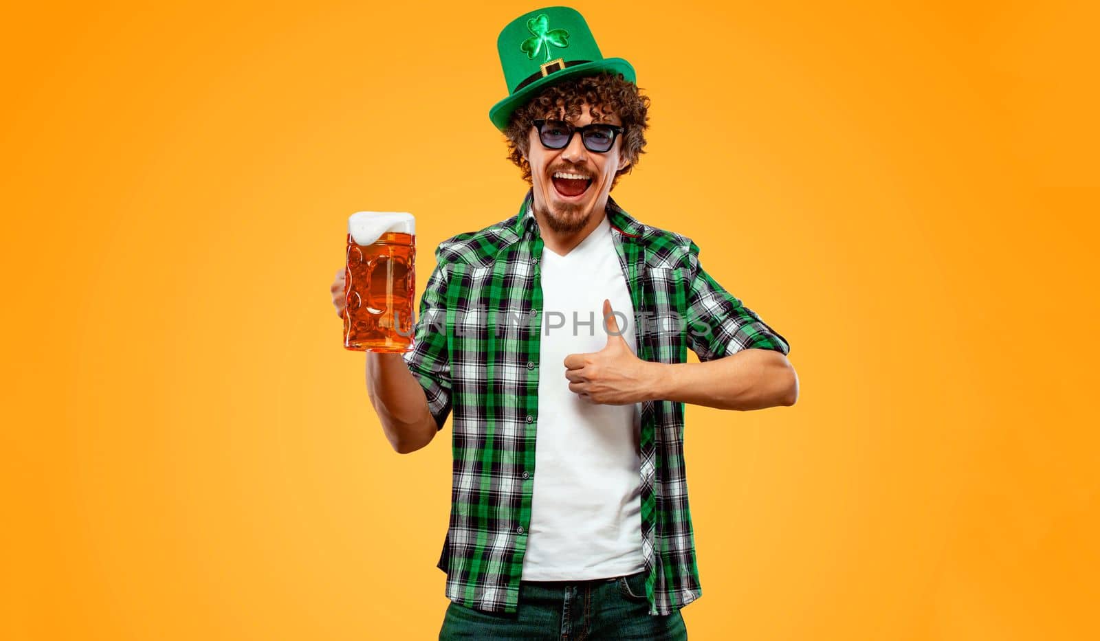 Saint Patrick Day. Young Oktoberfest man serving big beer mug with drink isolated on yellow background. Guy showing thumbs up sign with fingers. by MikeOrlov