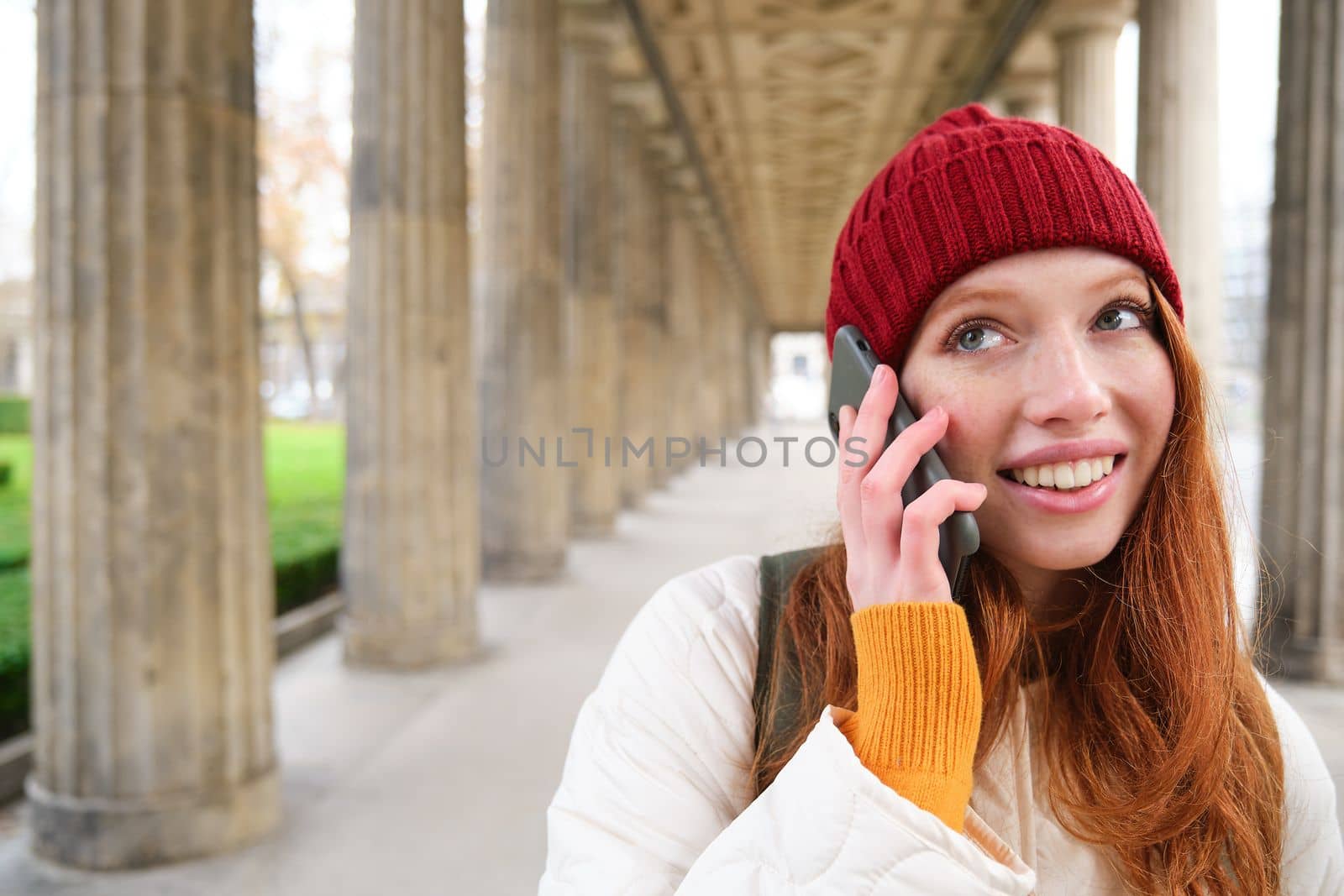 Smiling cute redhead woman makes a phone call, holds telephone near year, has mobile conversation, using smartphone on street.